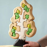 The Bumbu Toys Big Puzzle Birch Tree showing the green leaves side placed on a wooden surface in front of a blue background with an adult's hand holding on to the trunk.