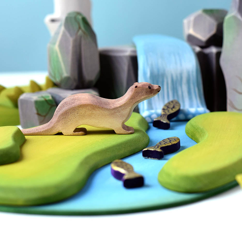 Close up of the Bumbu plastic-free wooden otter toy on a wooden waterfall scene next to some miniature fish toys