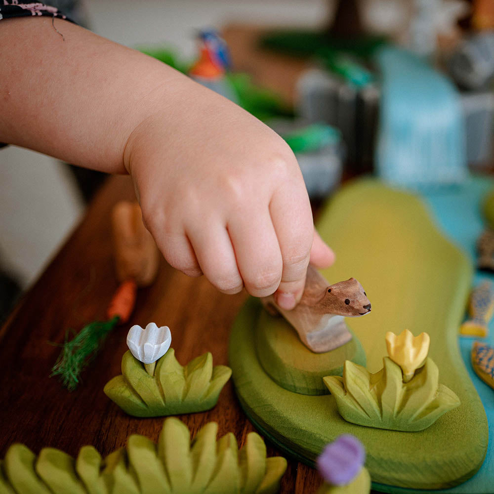 Close up of a hand moving a Bumbu wooden otter toy next to a Bumbu small wooden grass toy figure on a dark brown table