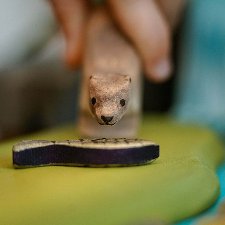 Close up of a childs hand holding the Bumbu solid wooden otter toy next to a miniature fish toy