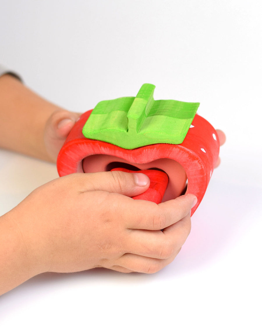 Hand slotting together the wooden pieces of the sustainable Bumbu stacking strawberry toy on a white background