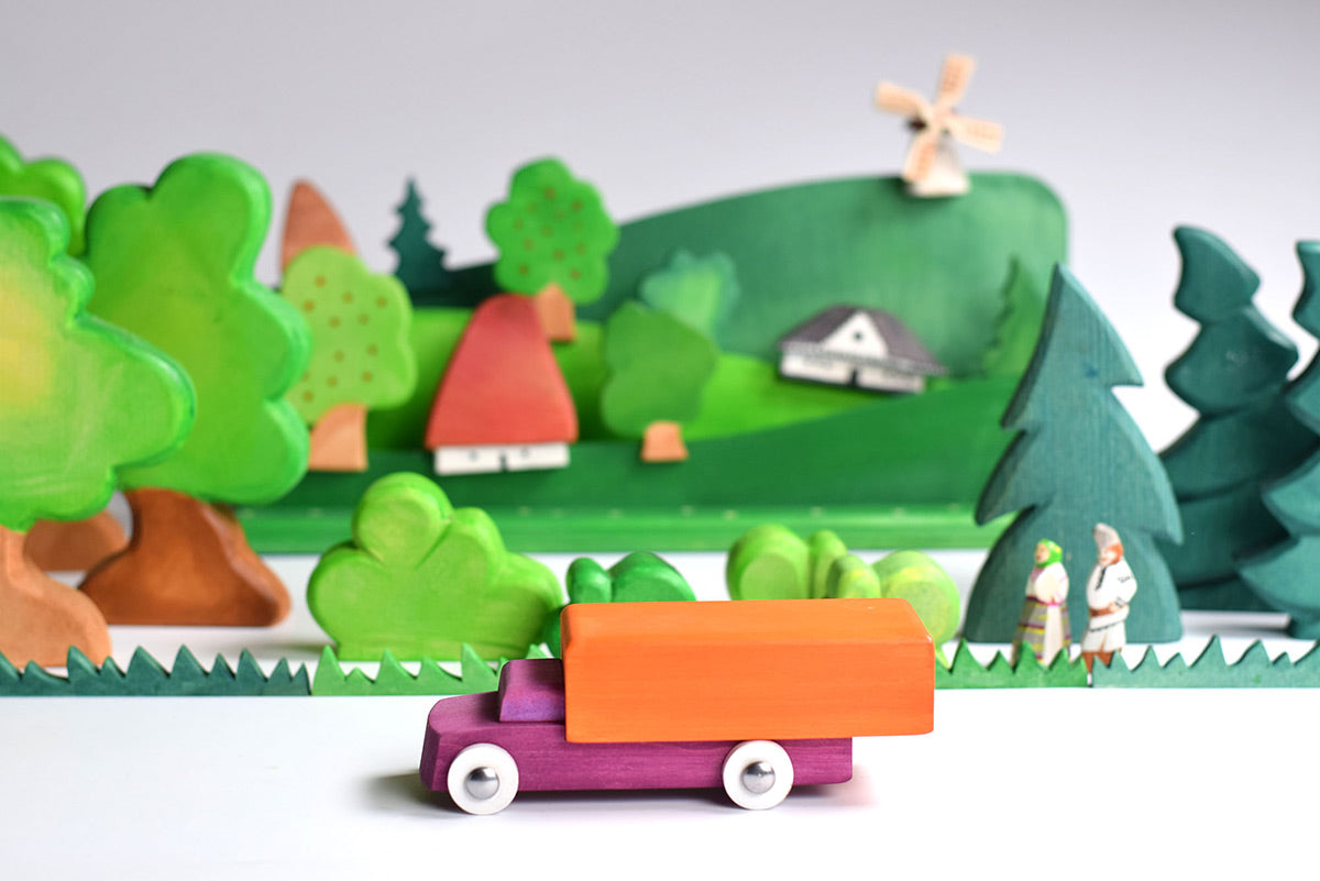 Bumbu wooden waldorf lorry toy on a white table in front of a green wooden toy landscape scene