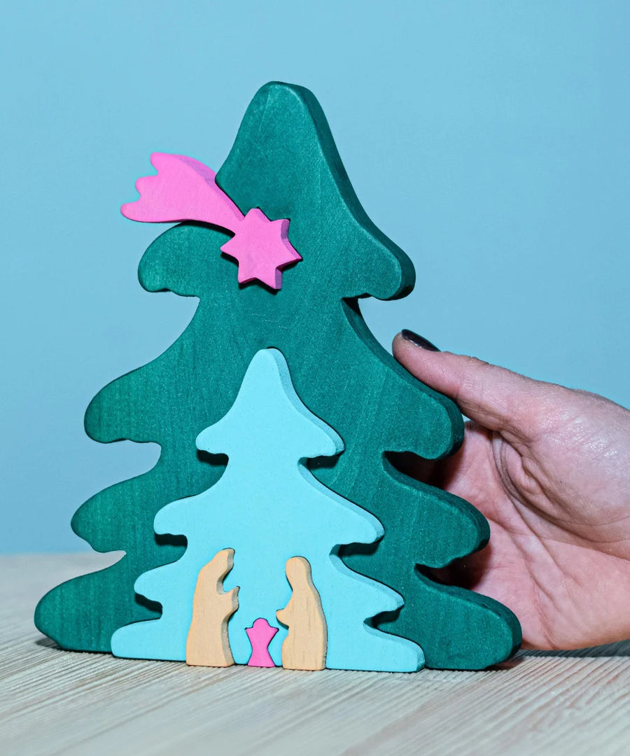 A Bumbu Christmas Tree Nativity Puzzle in a Fuchsia colour way placed on a wooden surface. An adult's hand can be seen on the side for scale.