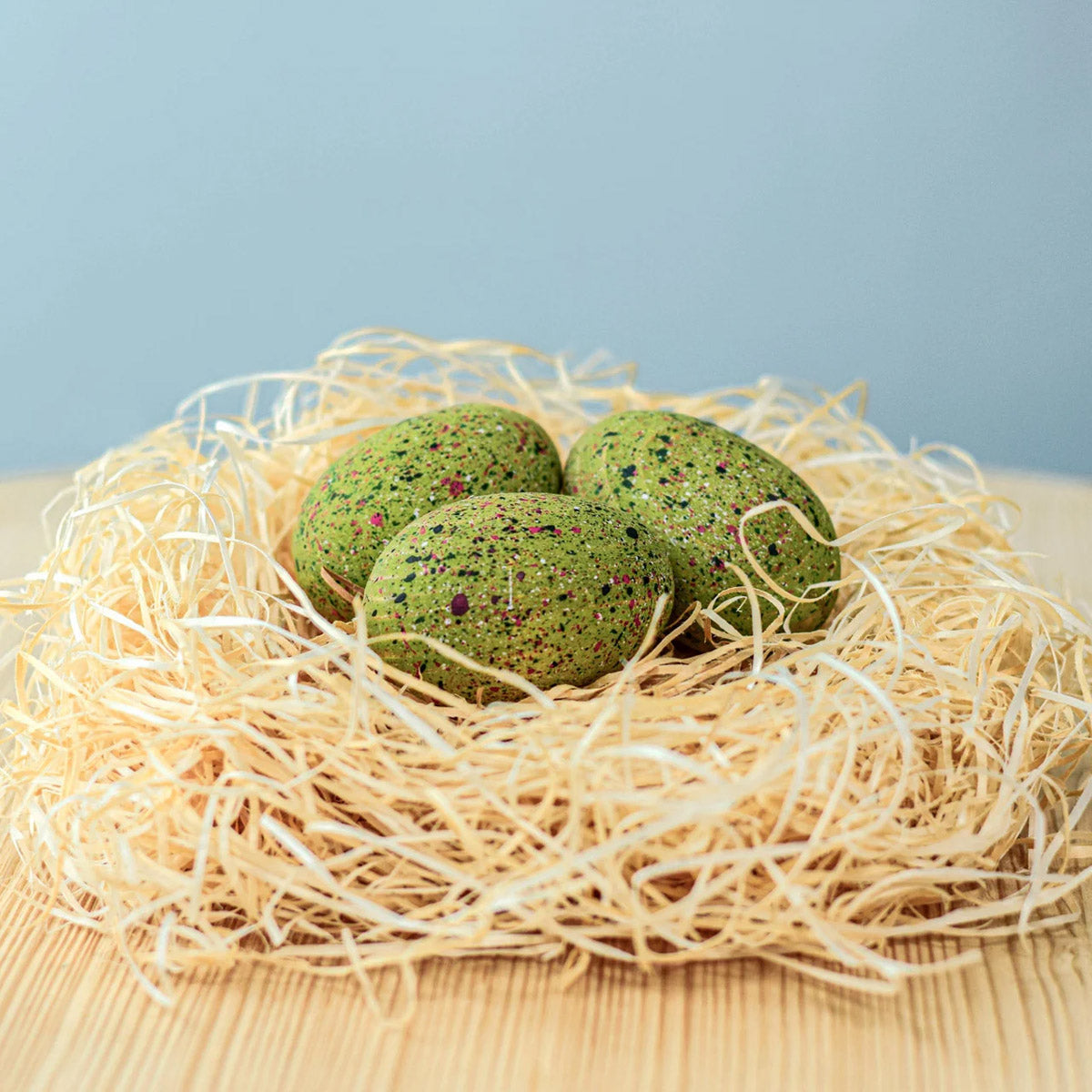 Bumbu Wooden T-Rex Eggs Set. Three speckled green dinosaur eggs sat in a nest. They are displayed on a wooden table and a blue background