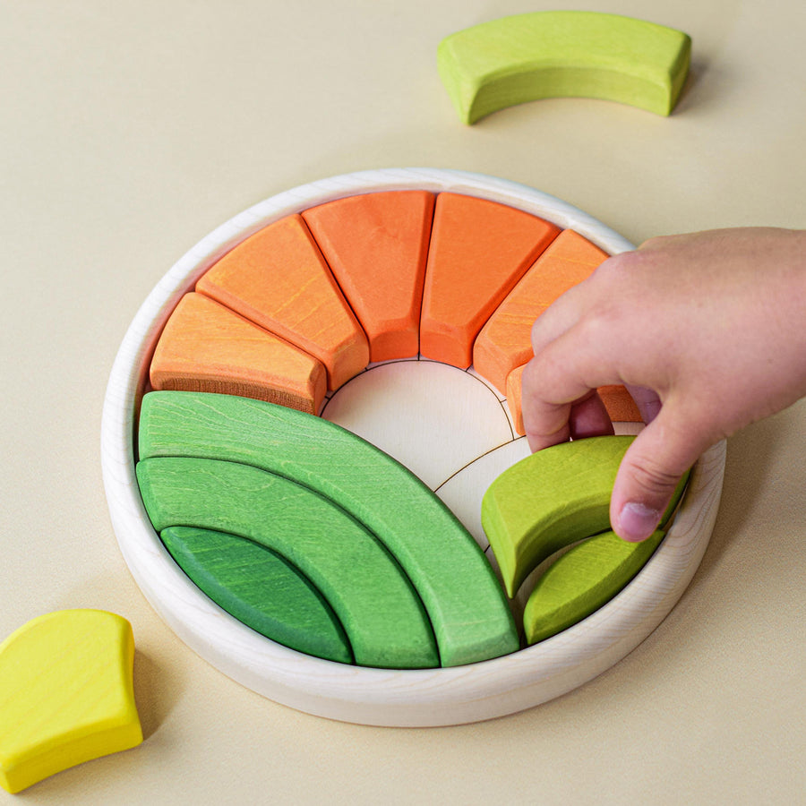 A circular-shaped wooden base is populated with colourful wooden blocks to create an image of the sun rising over two hills. There is a hand in the shot to denote the scale of the puzzle.