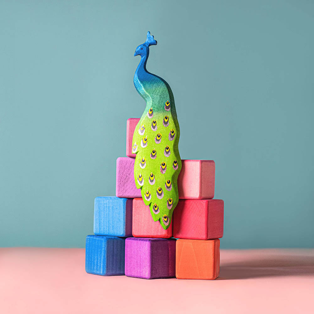 Close up of a Bumbu handmade wooden Peacock toy figure sat on some wooden toy blocks on a blue and pink background