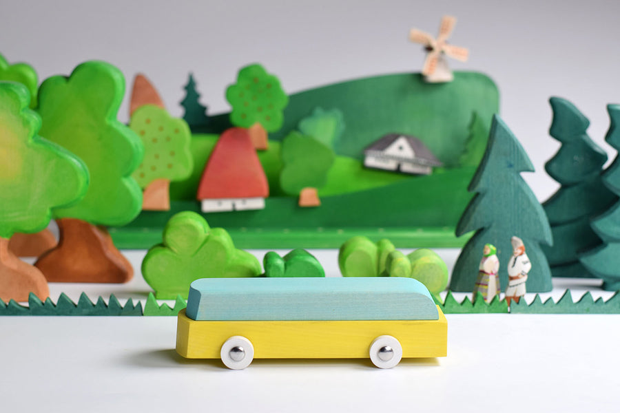 Bumbu waldorf car toy on a white table in front of a green wooden landscape scene