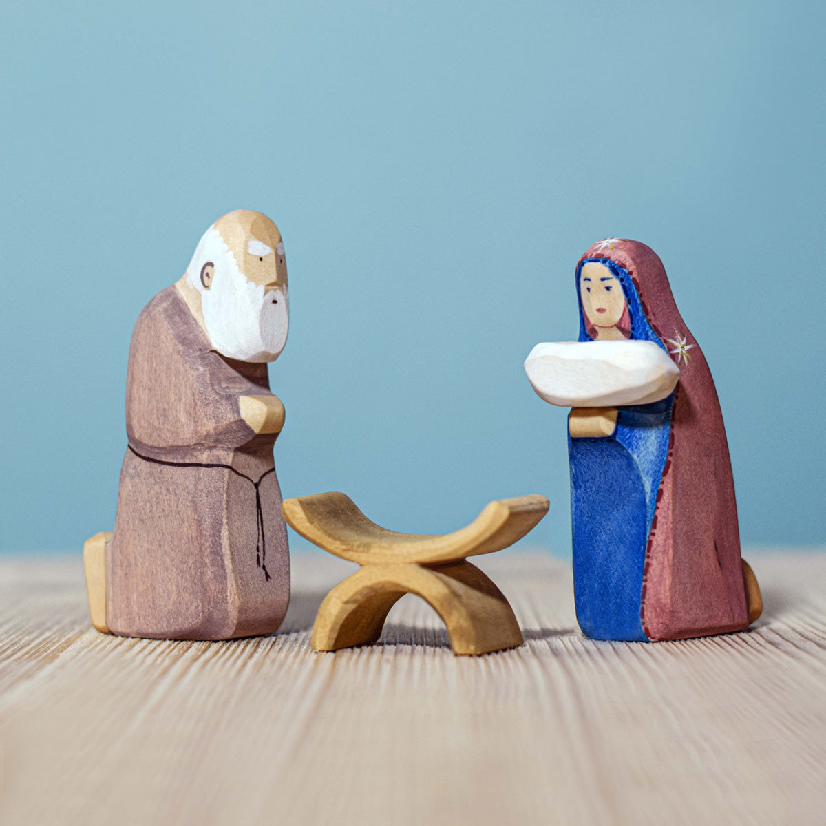 Bumbu Handmade Wooden Mary Figure kneeling down and holding the Bumbu Jesus. Mary is kneeling next to the Wooden Joseph figure and the wooden crib.