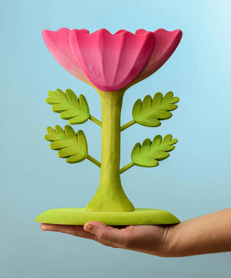 A person holding the Bumbu Wooden Large Pink Flower on their hand in front of a blue background.