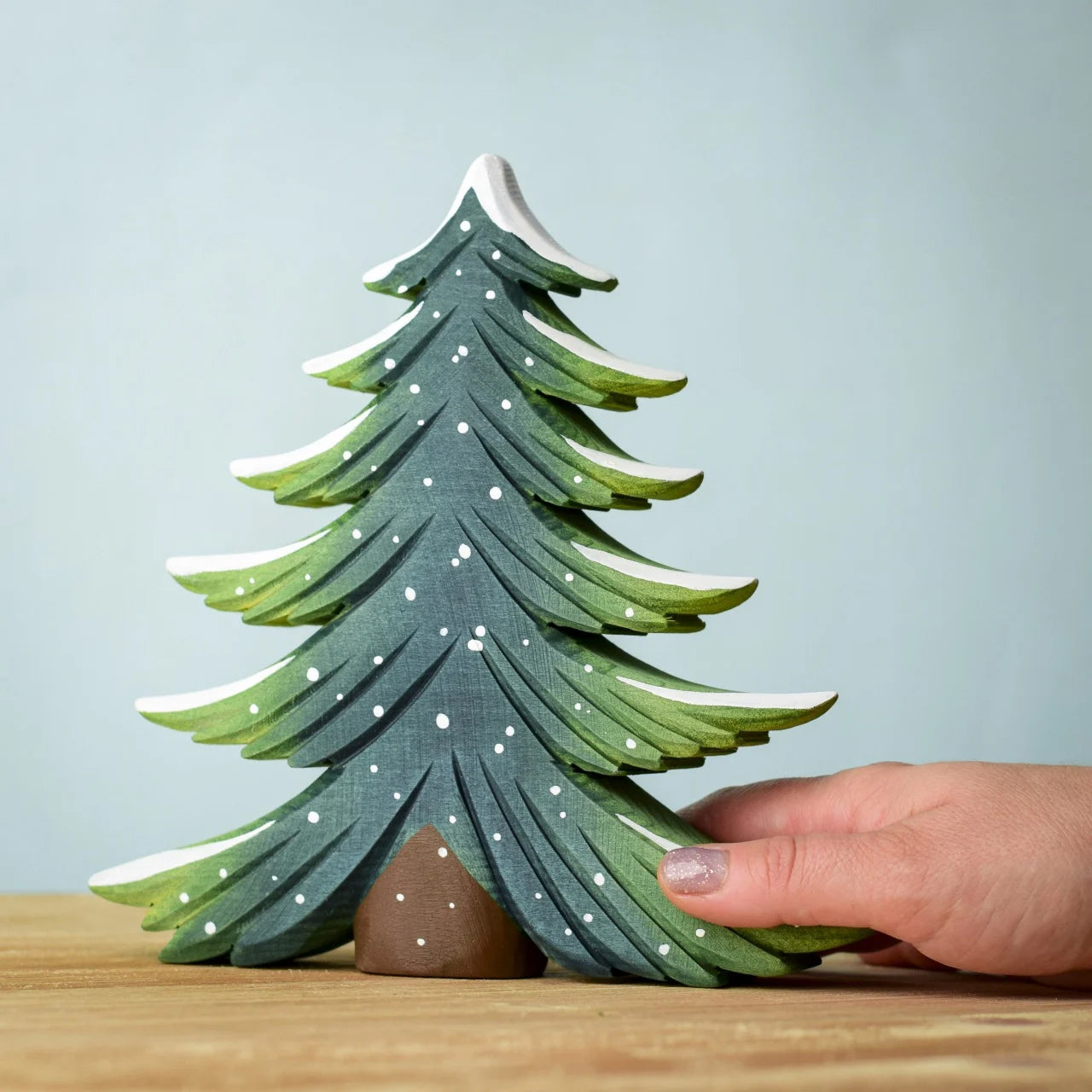 A large Bumbu Snowy Fir Tree placed on a wooden surface. An adult's hand can be seen in the side to show scale. 