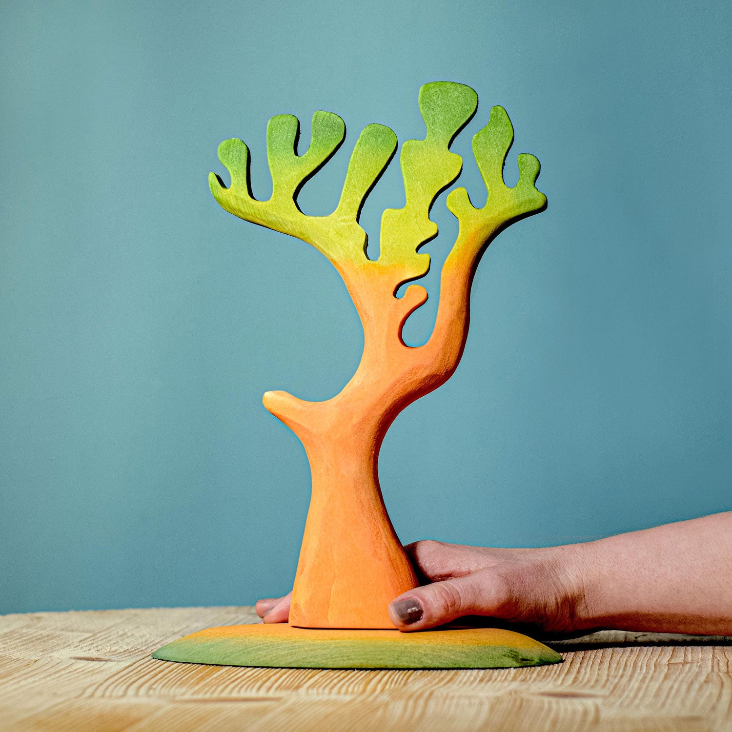 Adults hand holding the Bumbu Big Dino Tree placed on a wooden surface in front of a blue wall