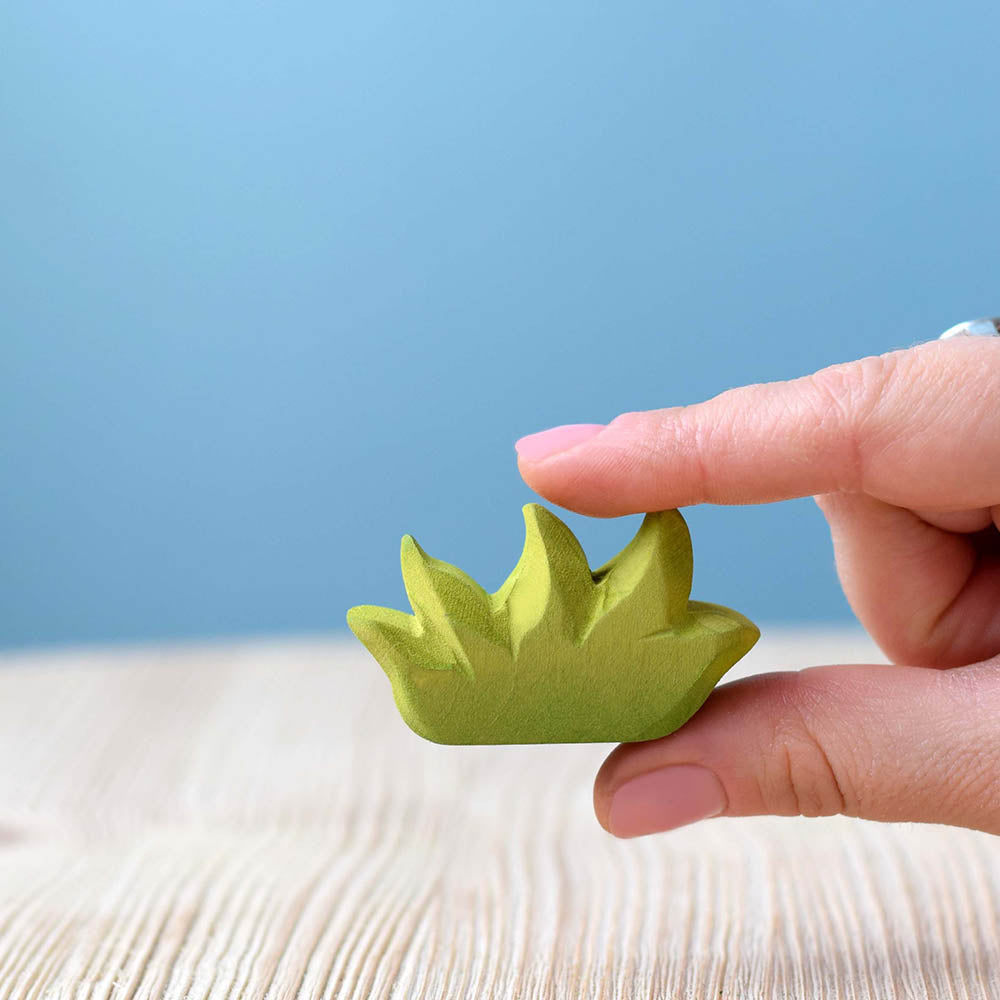 Close up of a hand holding the Bumbu handmade green wooden grass figure in front of a blue background