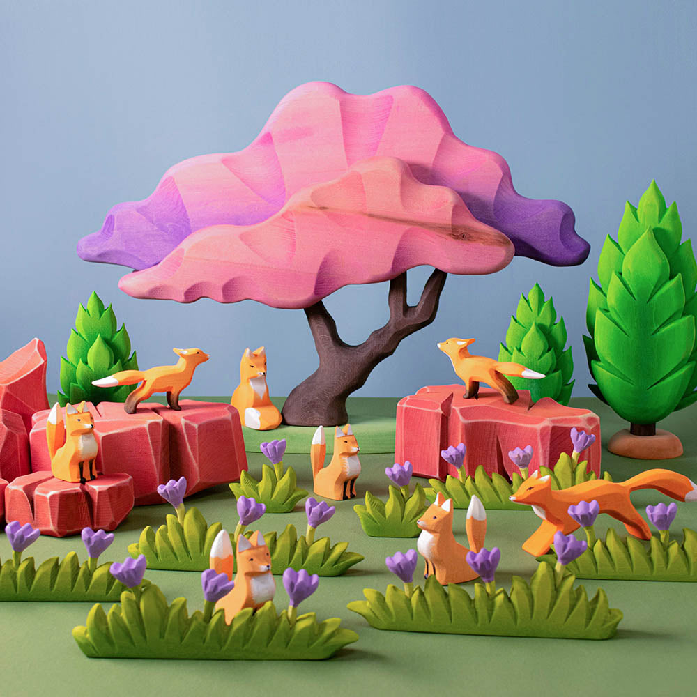 Bumbu plastic-free large grass figures with lilac flowers laid out on a green background next to some wooden fox figures and an Autumn maple tree toy in a small world scene