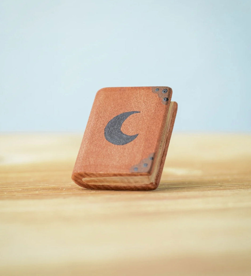 Bumbu Toys Small Book decorated with a black moon on the front. The book has dark brown corner plates and black dots. The book is propped up on a wooden surface.