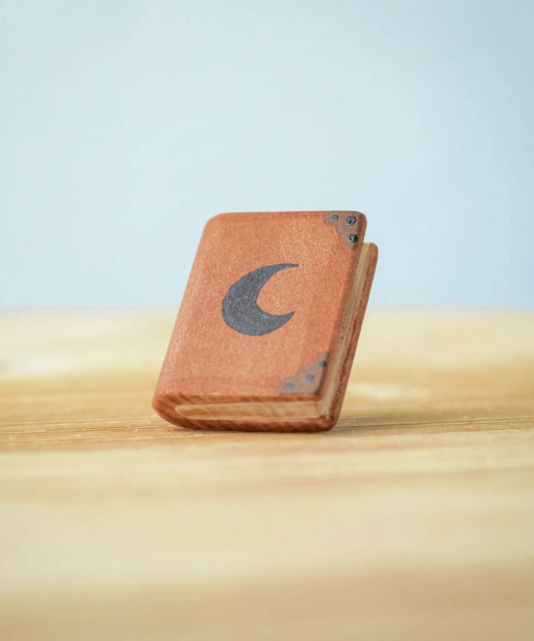 Bumbu Toys Small Book decorated with a black moon on the front. The book has dark brown corner plates and black dots. The book is propped up on a wooden surface.