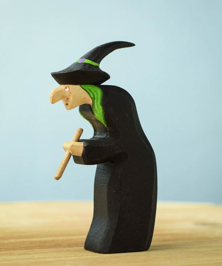 Bumbu Toys Brewing Witch wooden figure placed on a wooden surface. The witch has bright green hair and is wearing a black hat decorated with a painted bright green ribbon around the brim. The witch is clutching a wooden brewing stick in her hands. 