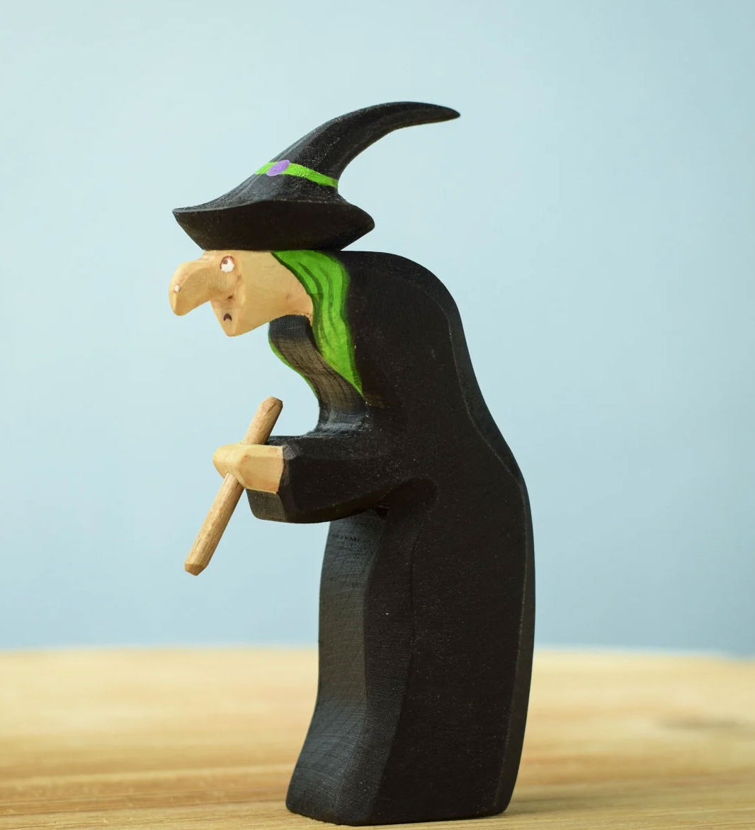 Bumbu Toys Brewing Witch wooden figure placed on a wooden surface. The witch has bright green hair and is wearing a black hat decorated with a painted bright green ribbon around the brim. The witch is clutching a wooden brewing stick in her hands. 