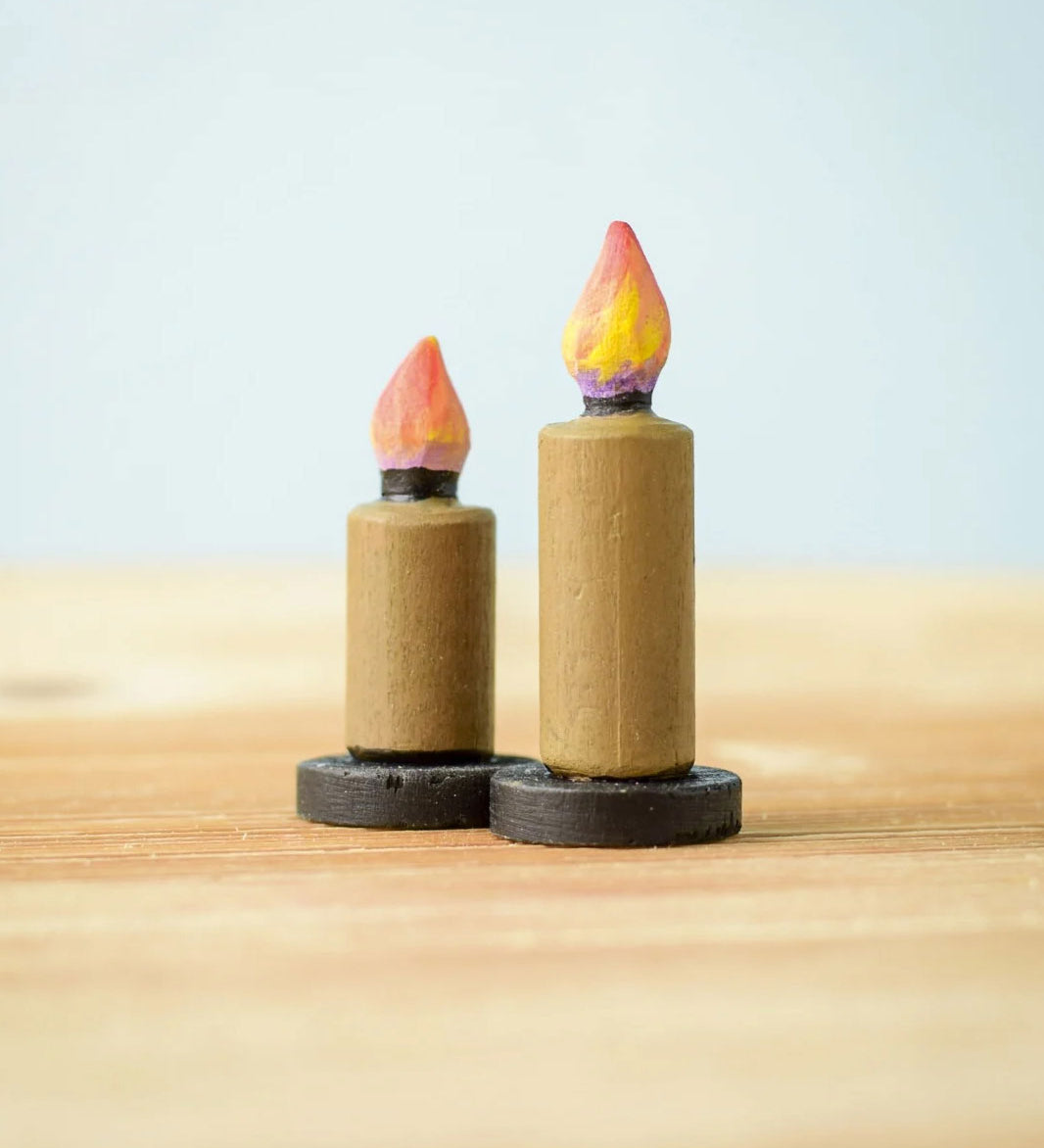 Bumbu Toys wooden Candle figures Set. The candles have a beige colour, on a black coloured base with a flame on the top. One candle is taller than the other. The candles are placed on a wooden surface. 