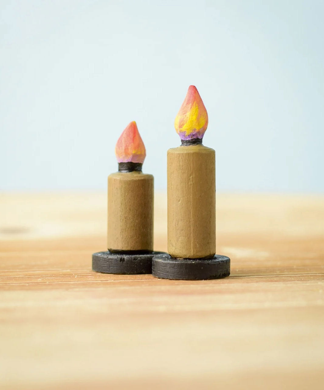 Bumbu Toys wooden Candle figures Set. The candles have a beige colour, on a black coloured base with a flame on the top. One candle is taller than the other. The candles are placed on a wooden surface. 