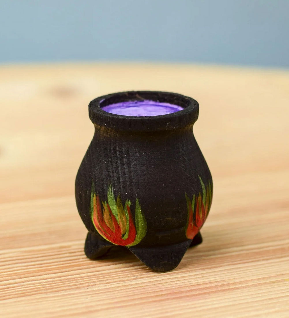 A Bumbu Wooden Witch's Cauldron. The cauldron is black on the outside and decorated with red, yellow and orange painted flames on the side. The top of the cauldron has a purple swirled effect decoration. The cauldron is placed on a wooden surface.