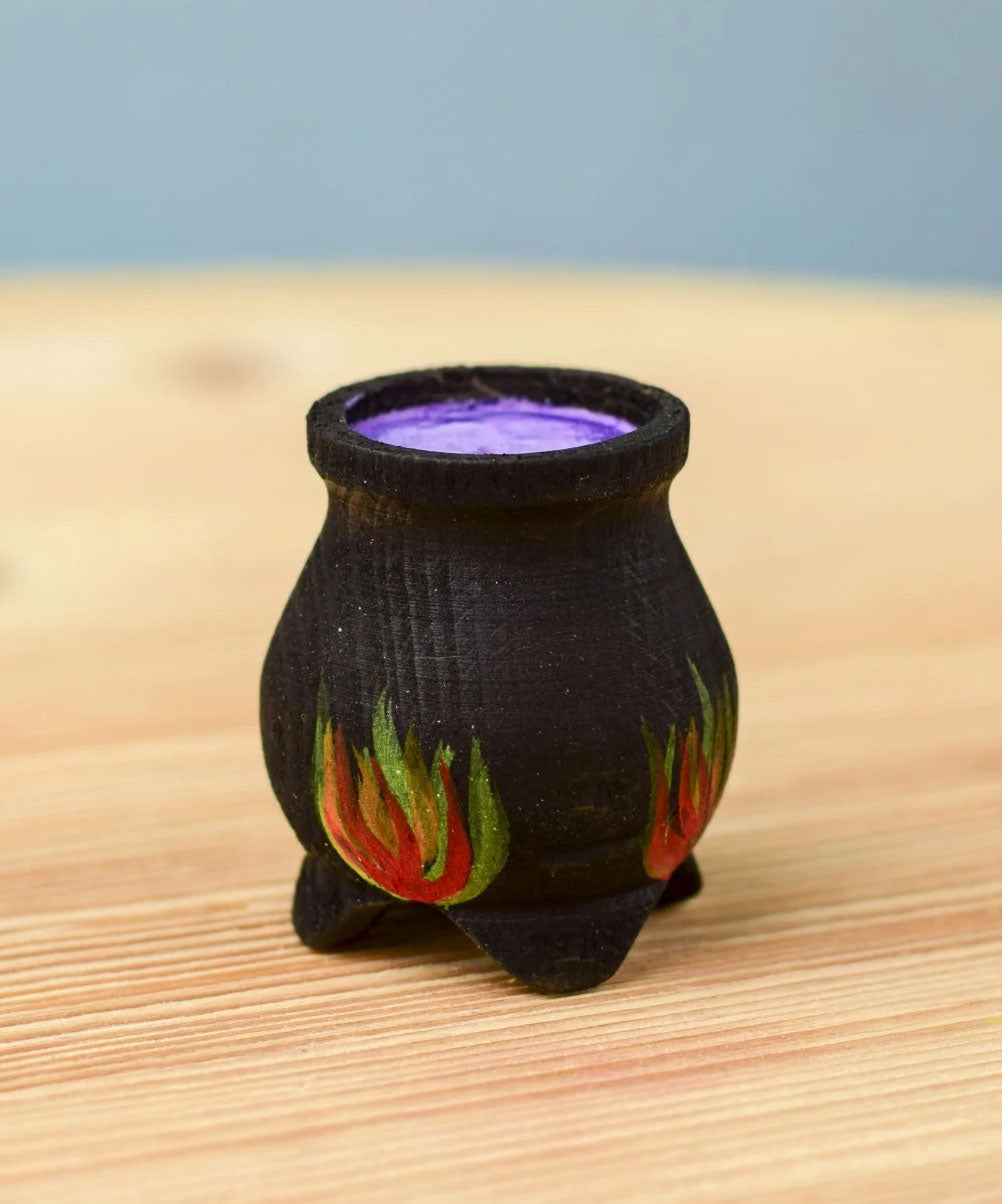 A Bumbu Wooden Witch's Cauldron. The cauldron is black on the outside and decorated with red, yellow and orange painted flames on the side. The top of the cauldron has a purple swirled effect decoration. The cauldron is placed on a wooden surface.