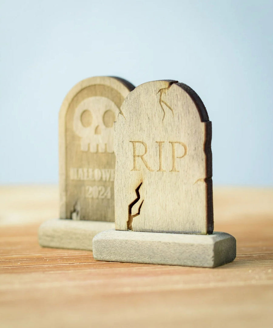 Bumbu Toys Gravestones Set. The set contains two headstones, one is decorated with RIP in large letters the other had a skull with Halloween 2024 written underneath. The Headstones are placed on a wooden surface.