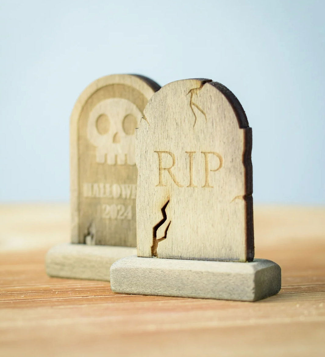 Bumbu Toys Gravestones Set. The set contains two headstones, one is decorated with RIP in large letters the other had a skull with Halloween 2024 written underneath. The Headstones are placed on a wooden surface.