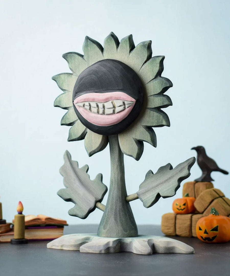 Bumbu Toys black wooden sunflower figure with a mouth with light pink lips and white teeth carved into it for a creepy Halloween vibe. The toy sunflower figure is placed on a wooden surface and surrounded by other Halloween themed wooden toys. 