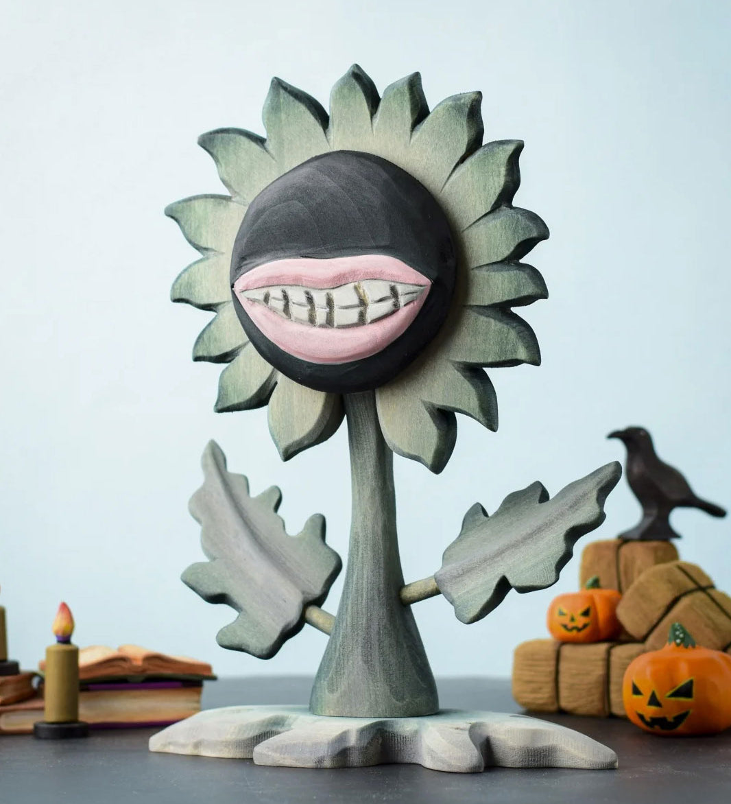 Bumbu Toys black wooden sunflower figure with a mouth with light pink lips and white teeth carved into it for a creepy Halloween vibe. The toy sunflower figure is placed on a wooden surface and surrounded by other Halloween themed wooden toys. 