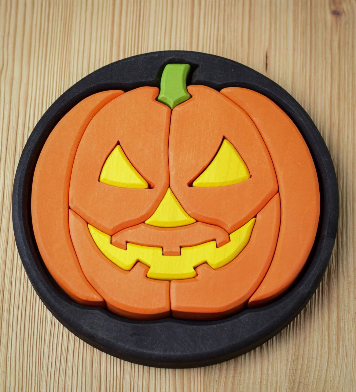 The Bumbu Halloween Pumpkin Round Wooden Puzzle pictured on it's black coloured base frame. The puzzle is on a wooden surface.