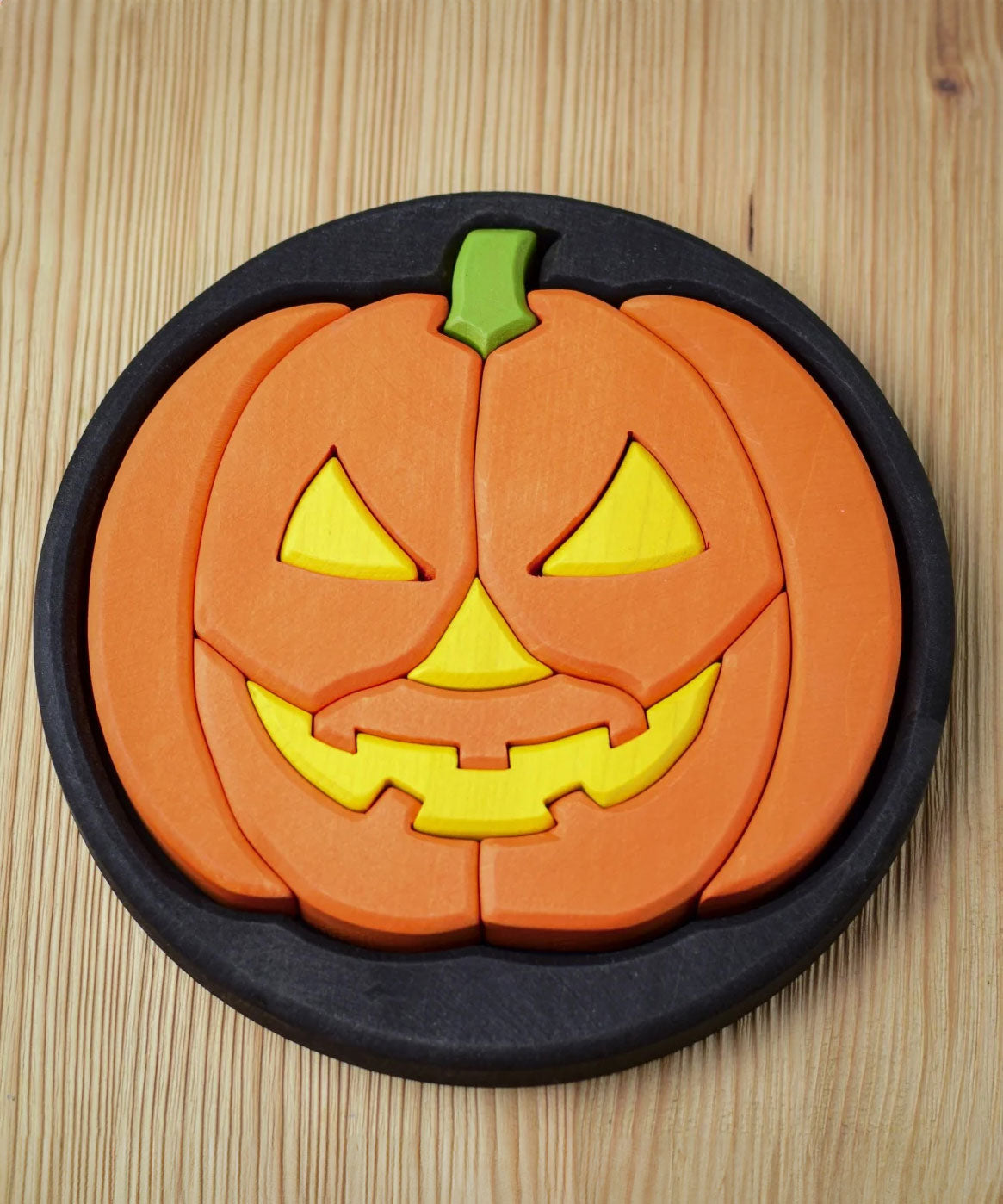 The Bumbu Halloween Pumpkin Round Wooden Puzzle pictured on it's black coloured base frame. The puzzle is on a wooden surface.