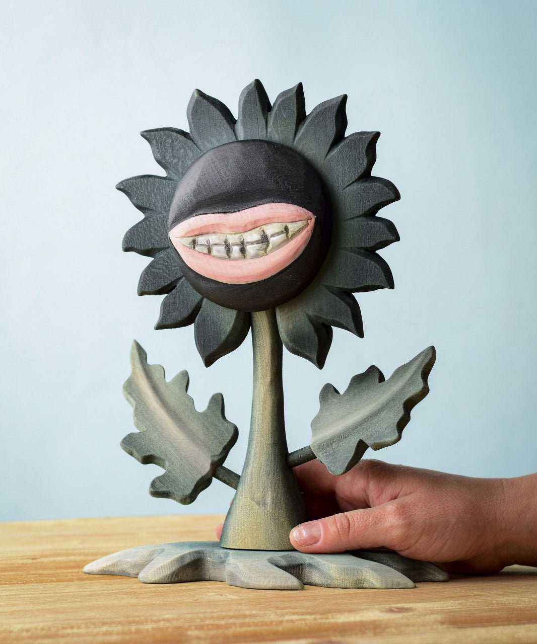 A hand reaching for the Bumbu Toys black wooden sunflower figure with a mouth with light pink lips and white teeth carved into it for a creepy Halloween vibe. The toy sunflower figure is placed on a wooden surface. 