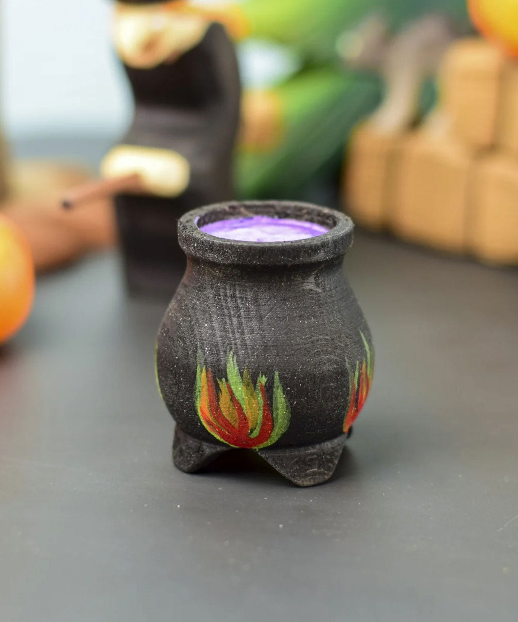 A Bumbu Wooden Witch's Cauldron. The cauldron is black on the outside and decorated with red, yellow and orange painted flames on the side. The top of the cauldron has a purple swirled effect decoration. The cauldron is placed at the front of a Halloween themed play scene, other wooden toys can be seen in the background. 