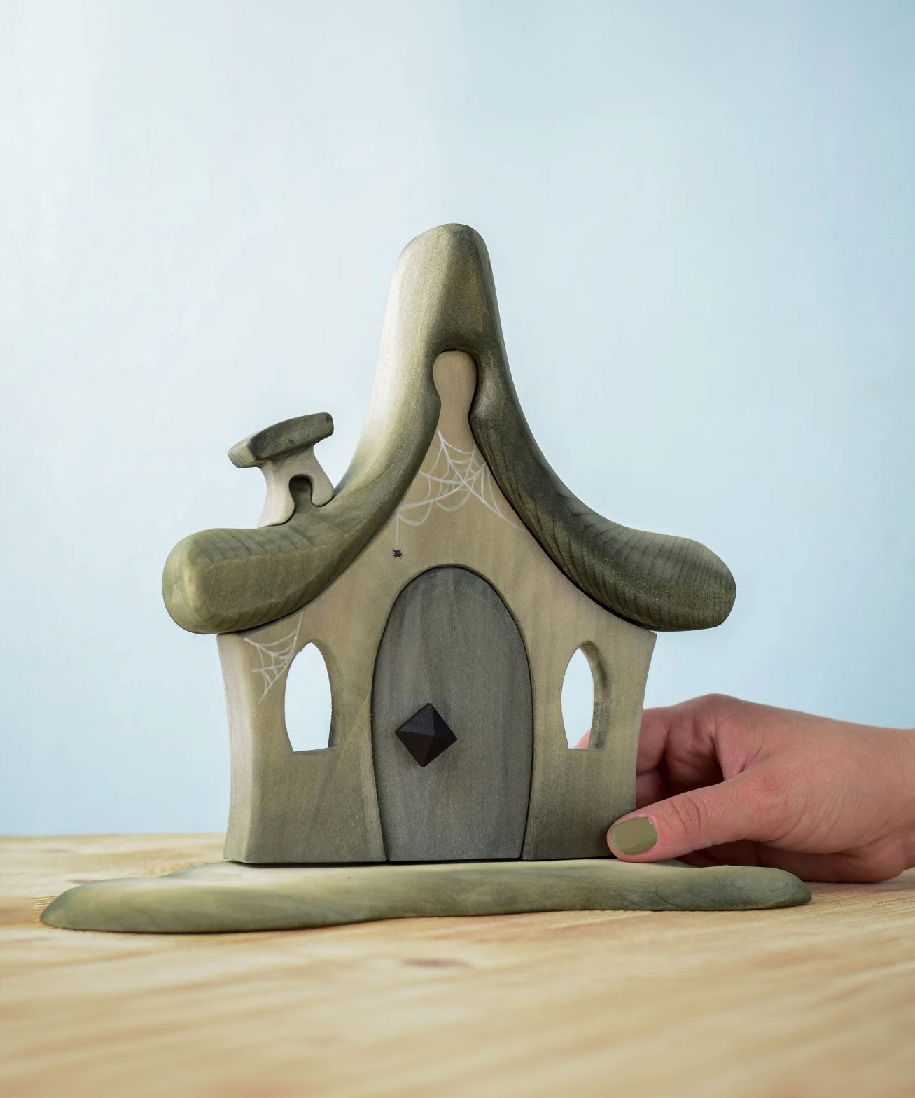 Bumbu Toys Witches house decorated with a Halloween theme. The house is grey in colour hand has cobwebs and a spider painted on. The House is placed on a wooden surface and an adult's hand can be seen holding one side. 