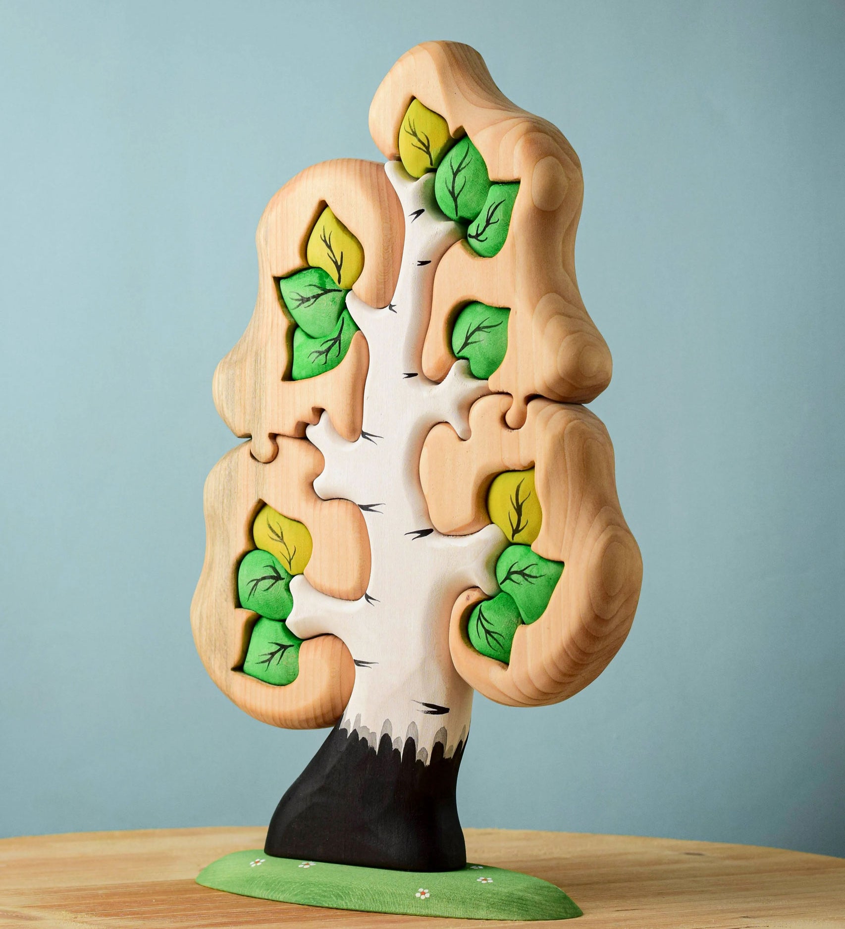 The Bumbu Toys Big Puzzle Birch Tree showing the green leaves side placed on a wooden surface in front of a blue background.