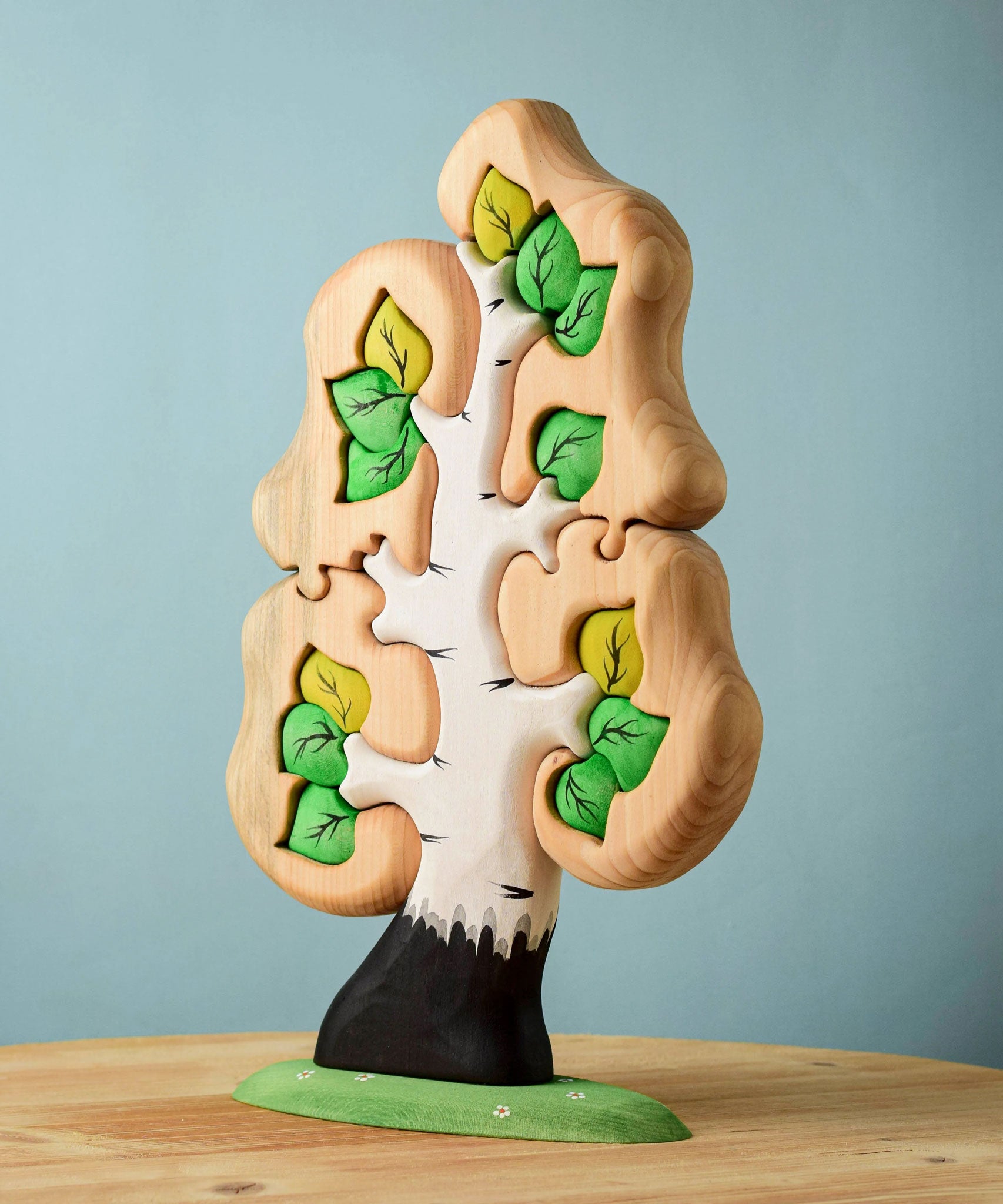 The Bumbu Toys Big Puzzle Birch Tree showing the green leaves side placed on a wooden surface in front of a blue background.