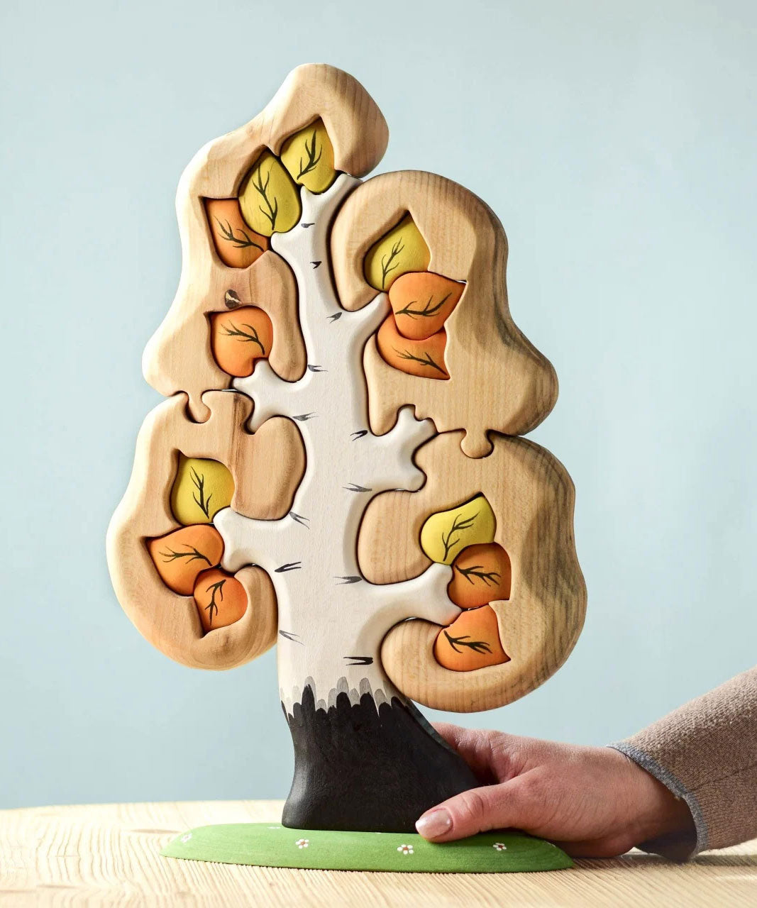 The Bumbu Toys Big Puzzle Birch Tree showing the orange and yellow leaves side placed on a wooden surface in front of a blue background with an adult's hand holding on to the trunk.