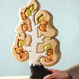 The Bumbu Toys Big Puzzle Birch Tree showing the orange and yellow leaves side placed on a wooden surface in front of a blue background with an adult's hand holding on to the trunk.