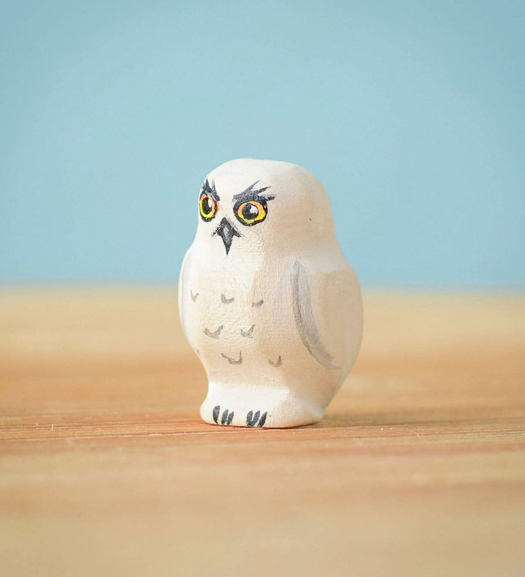 The Bumbu Toys Snowy Owl Chick placed on a wooden surface in front of a blue coloured background. 