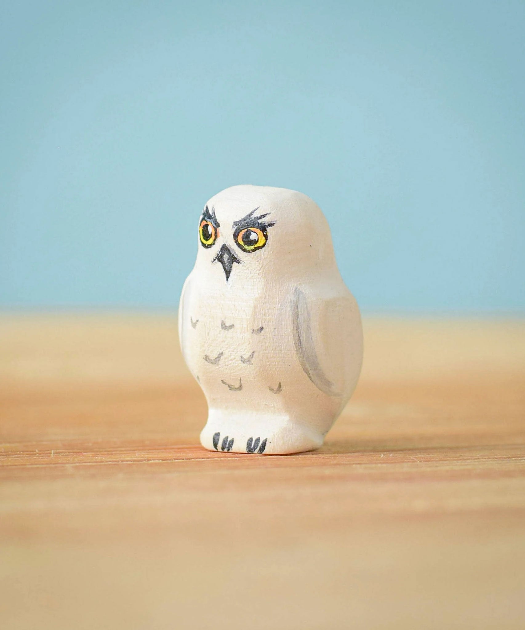 The Bumbu Toys Snowy Owl Chick placed on a wooden surface in front of a blue coloured background. 