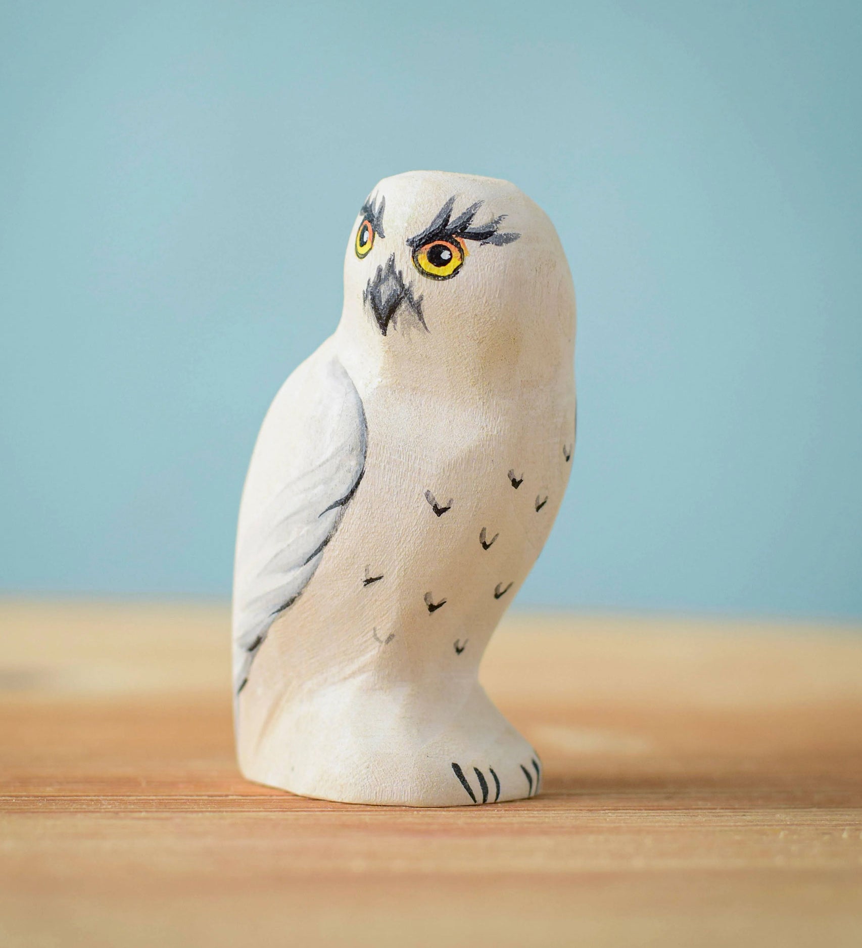 The Bumbu Toys Snowy Owl wooden toy figure placed on a wooden surface in front of a blue background.