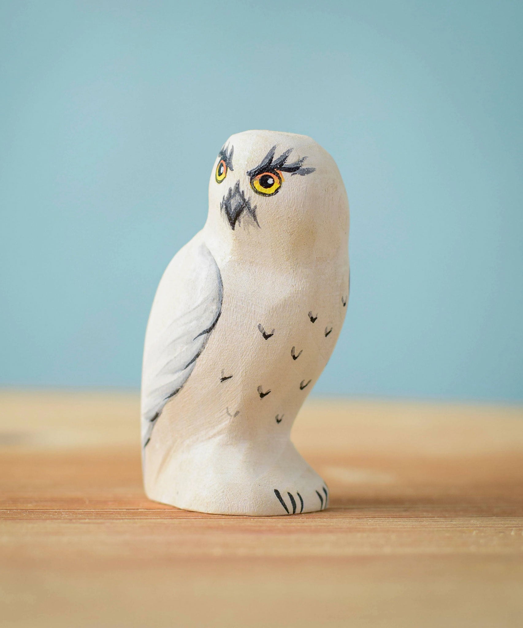 The Bumbu Toys Snowy Owl wooden toy figure placed on a wooden surface in front of a blue background.