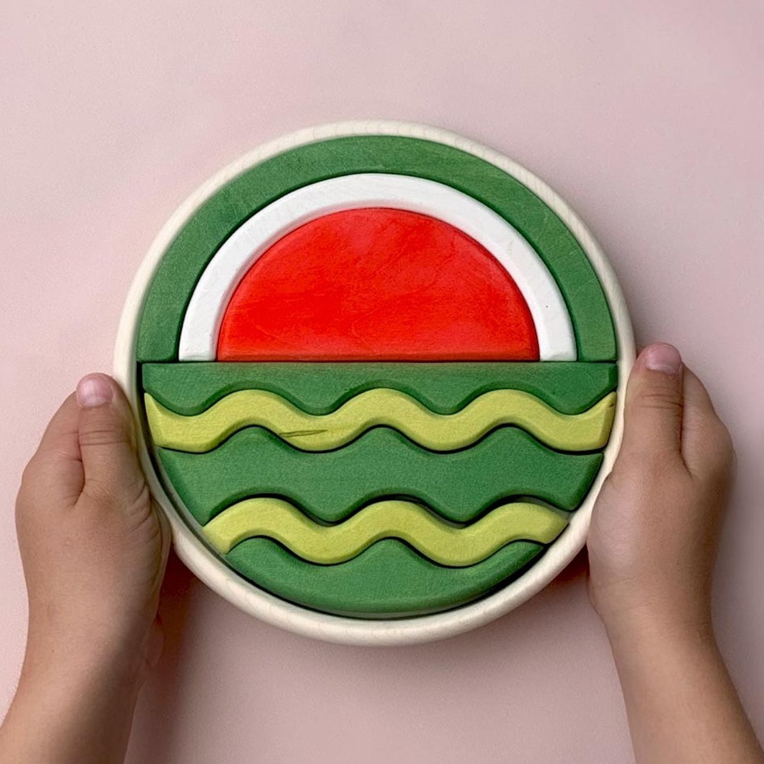 A round wooden puzzle depicting a watermelon in two halves. Different wooden shapes fit together to create the final puzzle. One half of the puzzle shows the inside of a watermelon whilst the other half shows the outside of a watermelon.