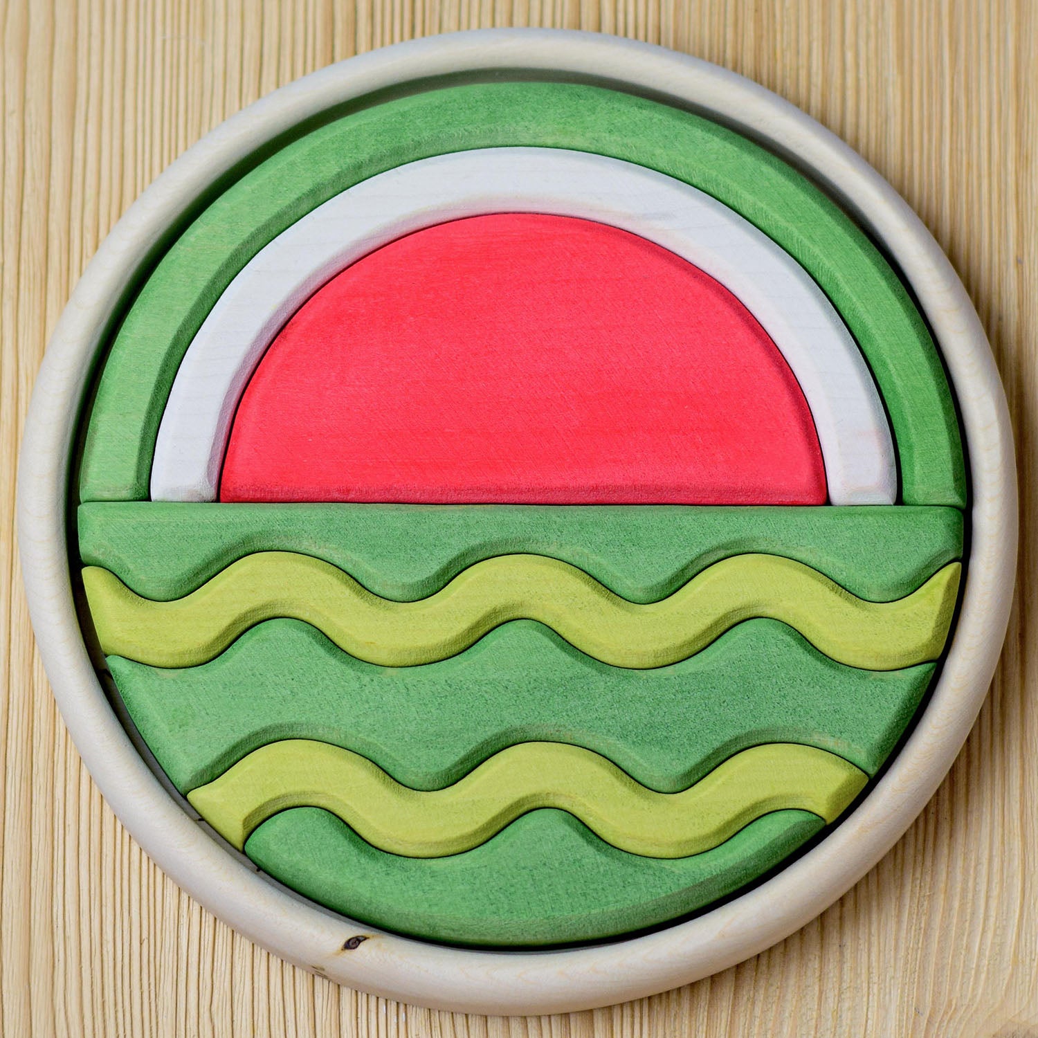 A round wooden puzzle depicting a watermelon in two halves. Different wooden shapes fit together to create the final puzzle. One half of the puzzle shows the inside of a watermelon whilst the other half shows the outside of a watermelon.