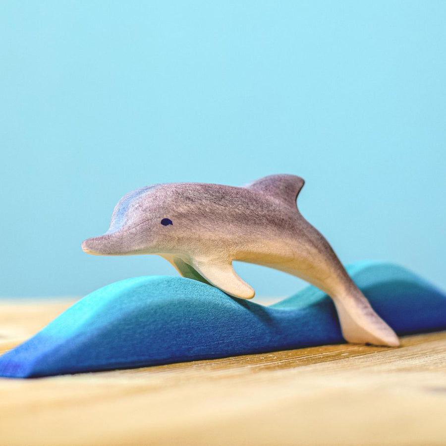 Bumbu Wooden Dolphin.  The toy is paired with a wave and sits on a wooden surface against a blue background.