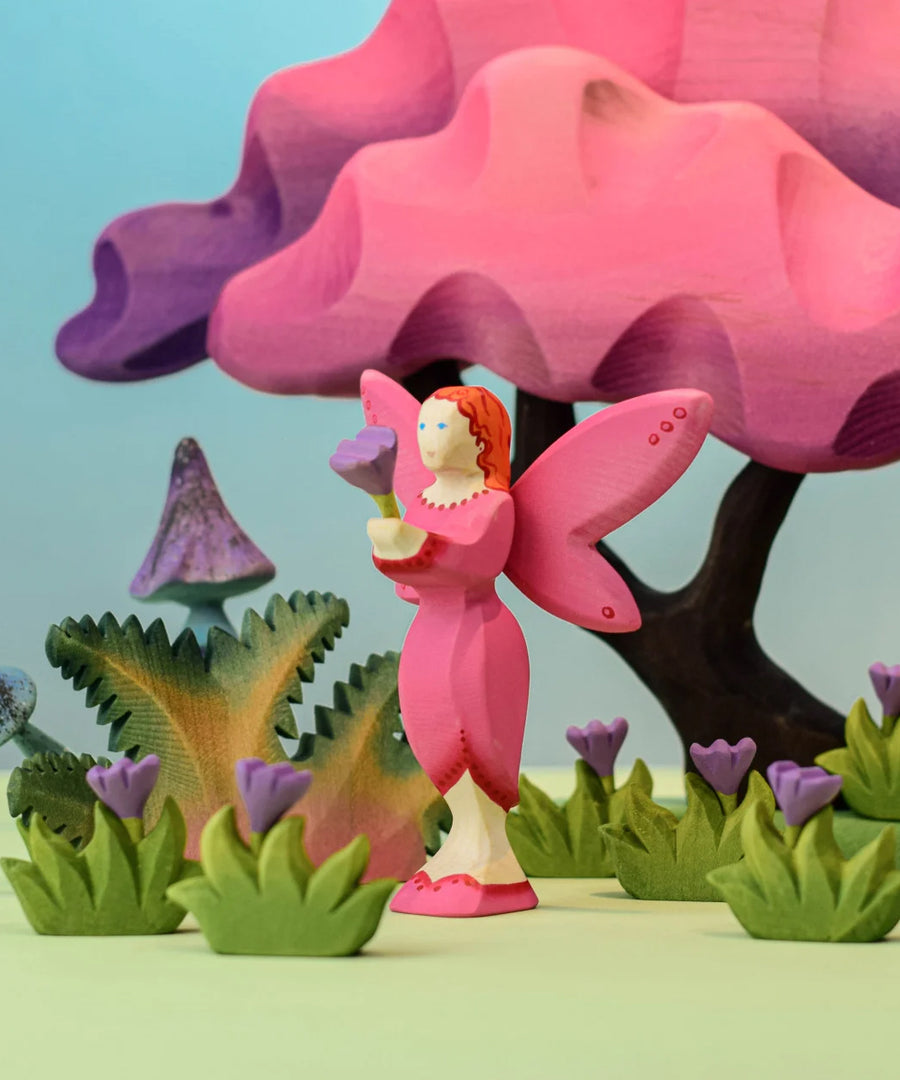 A Bumbu Wooden Blossom Fairy holding a small lilac flower in a small world play scene surronded by flowers with a Bumbu willow tree and  enchanted mushrooms in the background.