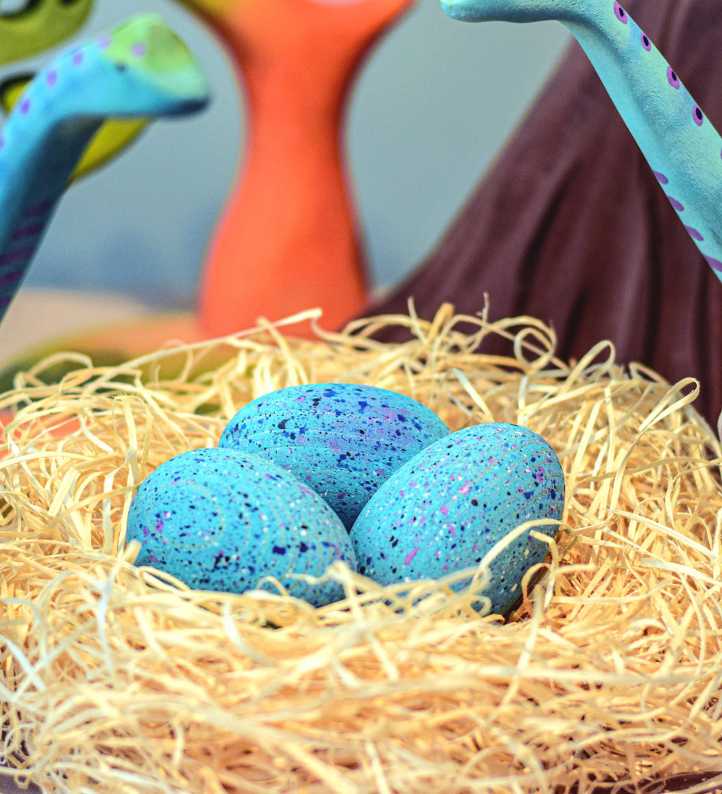 A set of Bumbu Wooden Brontosaurus Dinosaur Eggs in a nest. Dinosaurs and wooden trees can be seen in the background. 