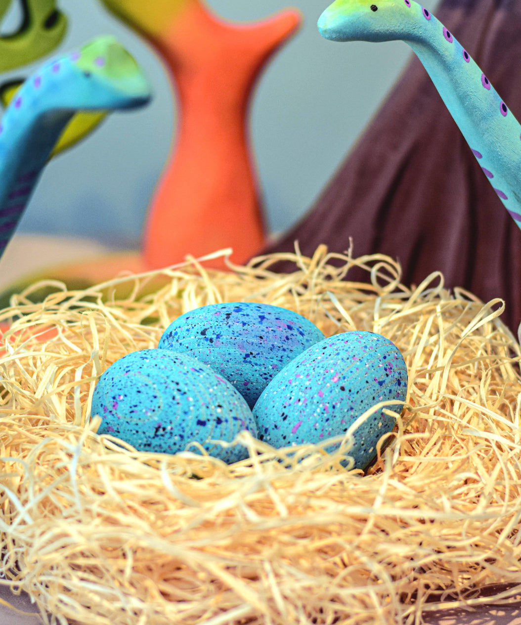 A set of Bumbu Wooden Brontosaurus Dinosaur Eggs in a nest. Dinosaurs and wooden trees can be seen in the background. 