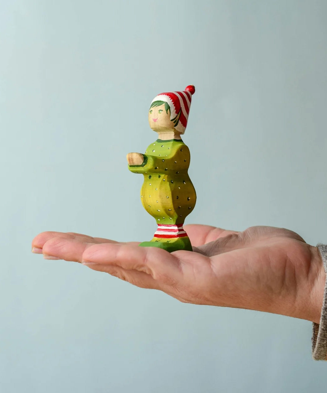 A hand holding the wooden Bumbu Santas elf wearing a red and white striped hat and a green outfit with its hands holding out to carry presents.