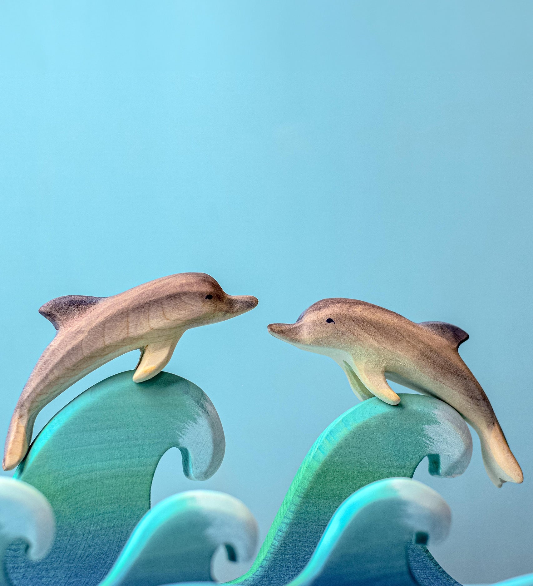 Two Bumbu Wooden Dolphins perched on waves in front of a blue background. 